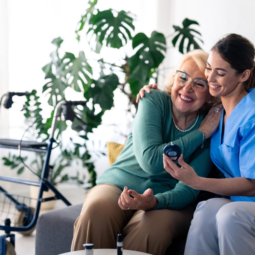 A smiling caucasian nurse in blue scrubs checks blood sugar levels for an elderly patient in a cozy apartment, using a glucometer while providing support and medical expertise.